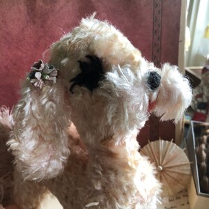May include: A white plush toy poodle with a black eye patch and a floral embellishment on its ear. The toy is in a sitting position and has a fluffy texture.