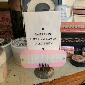May include: A white card with the text "IMITATION UPPER and LOWER FALSE TEETH" and a pink and white plastic set of false teeth on a metal stand.
