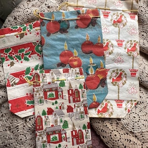 May include: Four vintage Christmas gift bags with different holiday designs. The bags are made of paper and have gold string ties. The designs include a red and white striped pattern with holly and birds, a blue pattern with red candles, a white pattern with red birds and pears, and a red and white pattern with houses and trees.