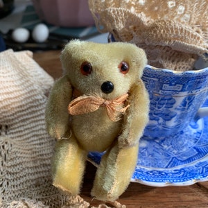 May include: A small, plush, light green teddy bear with brown eyes and a brown nose. The bear is wearing a light brown bow tie and is sitting on a blue and white teacup saucer.
