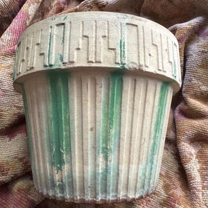 May include: A cylindrical terracotta plant pot featuring a cream-colored finish with vertical green painted stripes. The upper rim is decorated with a raised geometric pattern, and the exterior surface shows signs of weathered paint and texture.