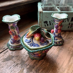 May include: A ceramic fruit-themed decorative set featuring a lidded container and two matching candle holders. The pieces are adorned with colorful fruit and leaf details, with a white interior and a blue and green trim. The set is placed on a weathered wooden surface.