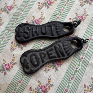 May include: Two black cast iron signs with a textured finish. One sign features the word SHUT in raised capital letters, and the other sign features the word OPEN in raised capital letters. Each sign has a circular hole at one end with a metal ring.