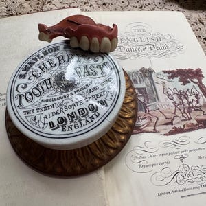 May include: An antique dental advertisement with a ceramic jar of "Tooth Cherry Paste" and a set of false teeth. The jar is on a decorative brass base, and the items rest on an open book with illustrations and text.