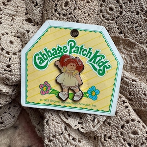 May include: A vintage Cabbage Patch Kids pin featuring a red-haired doll wearing a yellow dress and white shoes. The pin is on a yellow and green card with the text "Cabbage Patch Kids" and "©1983 Original Appalachian Artworks, Inc."