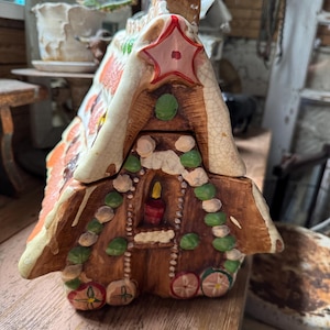 May include: A ceramic gingerbread house cookie jar with a red star on the roof. The house has white icing accents, green and white candies, and a candle in the window. The jar is brown with orange and white details.