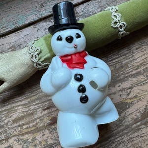 May include: A white snowman figurine with a black top hat, black coal eyes, and a red scarf. The snowman has three black buttons and a carrot nose. A festive winter decoration.
