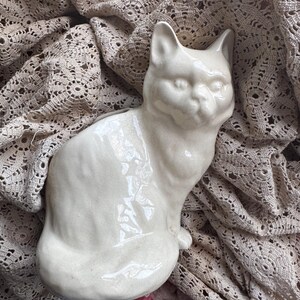 May include: A white ceramic cat figurine with a glossy finish. The cat is sitting with its head tilted slightly to the side. The figurine is on a crocheted fabric background.