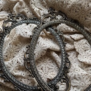 May include: Two antique oval picture frames with ornate, detailed bronze-colored metalwork. The frames are set against a backdrop of a vintage, cream-colored lace fabric, creating a classic, vintage aesthetic.