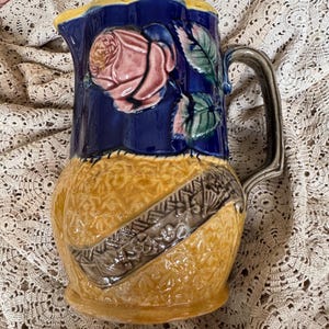 May include: A vintage ceramic pitcher with a rose motif. The pitcher features a blue upper section with a pink rose and green leaves. The lower section is yellow with a textured design and a gray band. The handle is gray.
