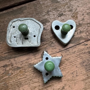 May include: Three vintage metal cookie cutters: an elephant, a heart, and a star. Each cutter has a green wooden handle. The cutters are displayed on a weathered, brown wooden surface.