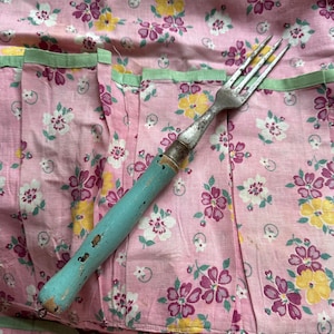 May include: A vintage metal fork with a weathered light blue painted wooden handle, featuring a three-pronged head, resting on a pink fabric background patterned with small white, yellow, and purple floral blossoms and green leaves.
