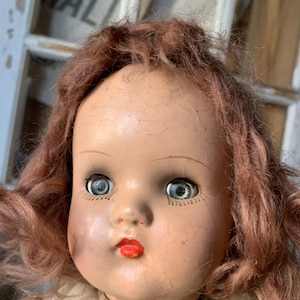 May include: A vintage doll with red hair and blue eyes. The doll's face is made of plastic and has a cracked appearance. The doll is wearing a white dress.