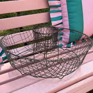 May include: An antique wire basket with a circular design and a smaller, central compartment. The basket is made of dark metal and sits on a pink bench. A striped green and pink pillow is in the background.