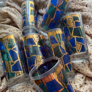 May include: A collection of six tall drinking glasses. Each glass features a mosaic design with blue and green geometric shapes, outlined in gold. The rims are also accented with gold. The glasses are arranged on a beige, textured surface.