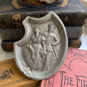 May include: A silver metal ashtray with a raised design of a couple in a vintage style. The man is wearing a military uniform and the woman is wearing a long dress. The ashtray is shaped like a horseshoe.