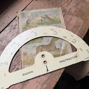 May include: A vintage protractor with radian markings and the word "PROTRACTOR" printed on it, resting on a worn, illustrated book. The book features animal illustrations and the letters "M" and "N". The protractor is off-white.