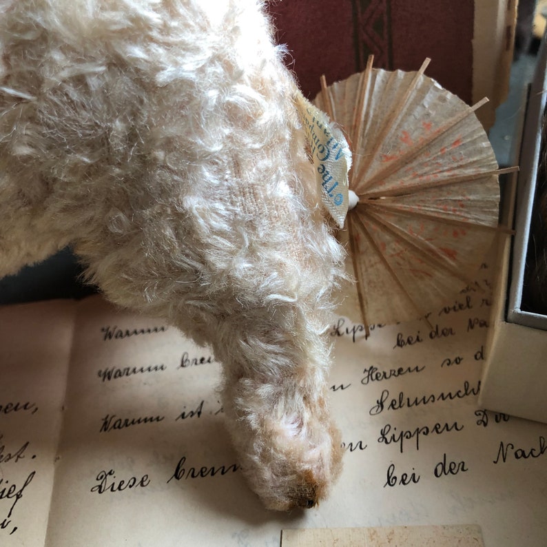 May include: A white, fluffy, vintage teddy bear with a tag that reads "The Gift" is standing on a page of a book with handwritten text. The teddy bear is looking down at the page.