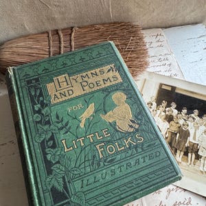 May include: An antique book titled "HYMNS AND POEMS FOR LITTLE FOLKS ILLUSTRATED" with a green cover and gold lettering. A vintage photograph and a broom are in the background. The book's cover features an illustration of a child and a bird.