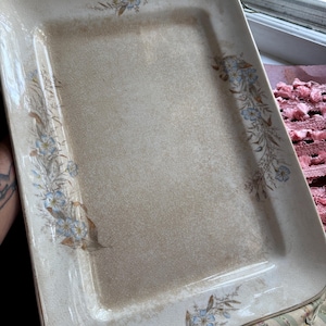 May include: A rectangular ceramic serving tray with a cream-colored base and a fine crackle glaze finish. The tray features delicate floral motifs in shades of blue and brown, arranged along the edges, with a smooth, slightly raised rim.