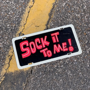 May include: A black and white license plate with the text "Sock It To Me!" in red.