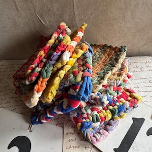 May include: A stack of colorful, braided pot holders in various hues, including red, yellow, blue, and green. The handmade kitchen accessories are made of fabric strips and feature a loop for hanging. The pot holders are a mix of woven and braided designs.