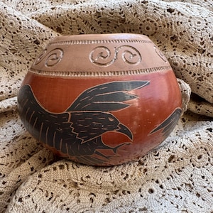 May include: A red clay pot with a black painted design of a bird in flight. The pot has a decorative rim with a swirl pattern.