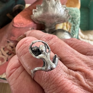 May include: A silver ring featuring a stylized figure in a dynamic pose. The figure appears to be leaping or dancing, with arms raised and legs extended. The ring has a smooth, polished finish and a minimalist design.