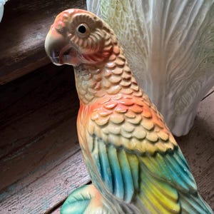 May include: A ceramic parrot figurine with a detailed design. The parrot features a brown head, orange and yellow feathers, and blue and green wings. It is perched on a branch with green leaves. The figurine has a glossy finish.
