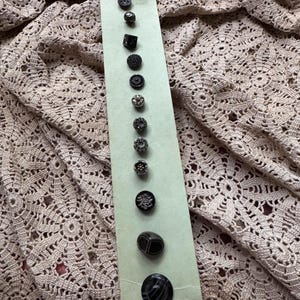 May include: A vertical light green card displaying twelve assorted black and dark gray buttons of varying shapes, sizes, and textures, arranged in a single column. The buttons feature diverse patterns, including floral, geometric, and smooth circular designs.