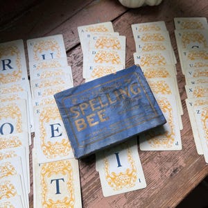 May include: Vintage spelling bee game set with a blue box and letter cards. The box reads "Spelling Bee" in gold lettering. The cards have ornate gold borders and individual letters. The set is arranged on a weathered wooden surface.