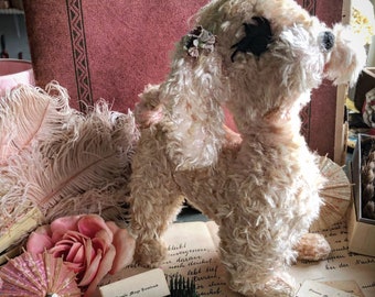 May include: A white, fluffy, vintage toy dog with black eyes and a pink bow. The dog is sitting on a book with a pink feather boa and a pink rose.