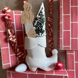 May include: A white ceramic boot with a red and white candy cane garland, a small artificial Christmas tree, and a wooden doll figure inside. The boot is sitting on a pink brick surface.