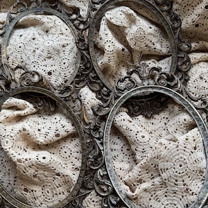 May include: Four antique oval picture frames with ornate, detailed scrollwork. The frames are a tarnished silver color, and the empty centers are visible. The frames are arranged on a cream-colored, crocheted fabric.