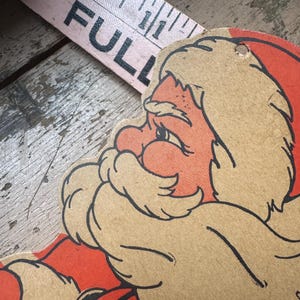 May include: A vintage cardboard cutout of Santa Claus with a red hat and white beard. The cutout is partially torn and has a hole in the top right corner.