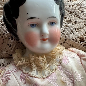 May include: Antique porcelain doll with a serene expression, blue eyes, and rosy cheeks. The doll has black hair and wears a pink dress with a lace collar. The dress has a floral pattern.
