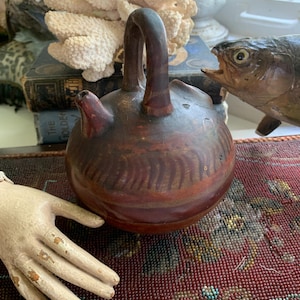 May include: A small, brown, ceramic pitcher with a handle and a spout. The pitcher has a red and brown painted design. The pitcher is sitting on a beaded surface.
