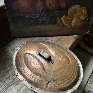 May include: Oval copper pot with a lid featuring embossed fish designs. The pot has a handle and is set on a wooden surface. The background includes a painting of oranges in a basket.