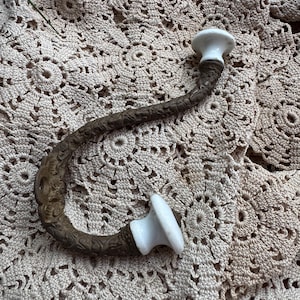 May include: A curved metal wall hook with a textured, dark bronze-toned finish and two smooth, white ceramic knobs at each end. The hook features a mounting plate with a visible screw hole, designed for hanging items on a vertical surface.