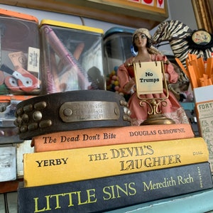 May include: A display featuring vintage books with titles like "The Dead Don't Bite" and "Little Sins." A studded leather collar sits atop the books. A sign that says "No Trumps" is displayed near a doll. A vintage tickets sign is in the background.