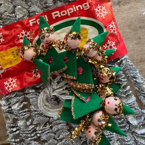 May include: Vintage Christmas elf ornaments with green felt bodies, gold trim, and painted faces. The elves are attached to a red and white package labeled "Foil Roping". The ornaments are on a silver foil surface.