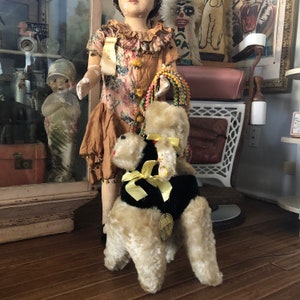 May include: A vintage plush toy poodle wearing a black velvet jacket with a yellow bow. The poodle is standing on a wooden floor. The toy is in excellent condition and has a tag that reads "First Pets".