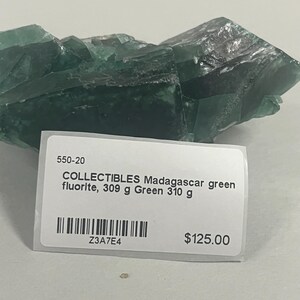 May include: A raw green fluorite crystal specimen from Madagascar with a white rectangular label attached. The label displays the text 550-20, COLLECTIBLES Madagascar green fluorite, 309 g Green 310 g, a barcode, Z3A7E4, and $125.00.