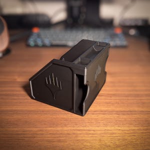 May include: A black, 3D-printed card holder with the Magic: The Gathering symbol. The holder has multiple compartments and angled sides. It sits on a wooden desk, with a keyboard and speaker in the background.