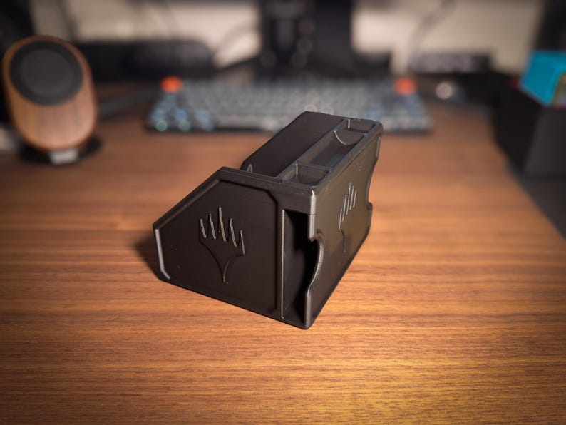 May include: A black, 3D-printed card holder with the Magic: The Gathering symbol. The holder has multiple compartments and angled sides. It sits on a wooden desk, with a keyboard and speaker in the background.