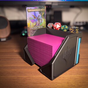 May include: A black card holder with a stack of pink cards, a Magic: The Gathering card, and several dice. The card holder has a blue accent and the Magic: The Gathering symbol. The dice are red, green, and black.