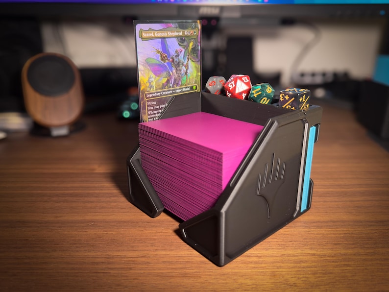 May include: A black card holder with a stack of pink cards, a Magic: The Gathering card, and several dice. The card holder has a blue accent and the Magic: The Gathering symbol. The dice are red, green, and black.
