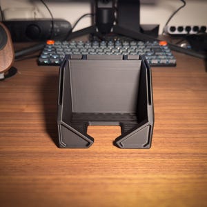 May include: A dark gray, 3D-printed desk organizer with a rectangular shape and angled sides. The organizer has a slot at the bottom and is designed to hold items. The product is sitting on a wooden desk.