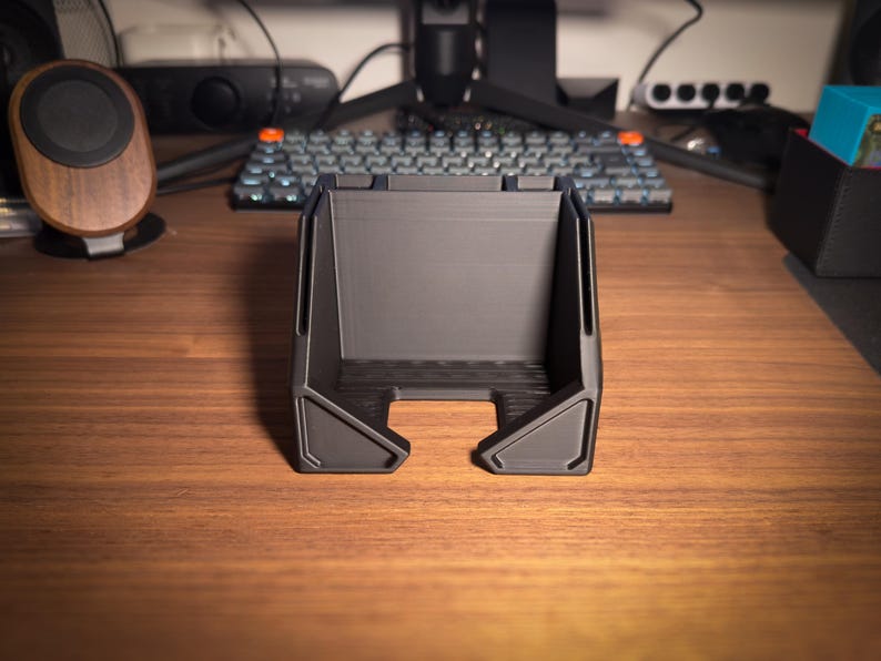 May include: A dark gray, 3D-printed desk organizer with a rectangular shape and angled sides. The organizer has a slot at the bottom and is designed to hold items. The product is sitting on a wooden desk.