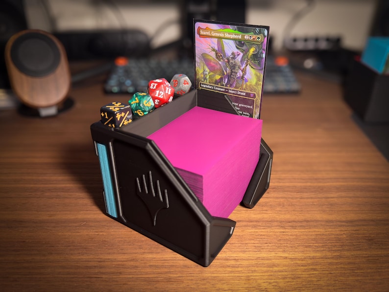 May include: Black and teal card holder with a stack of pink cards, dice, and a Magic: The Gathering card. The holder has a stylized design with a logo on the front. The dice are red, green, and gray.
